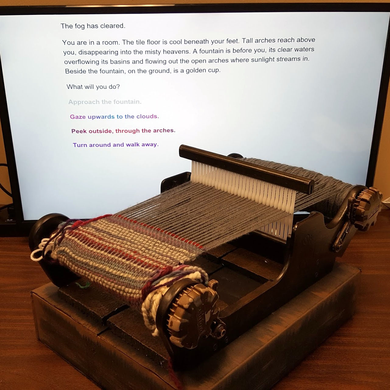 A handheld loom with in-progress work sits in front of a computer screen with a text choose-your-own-adventure game.